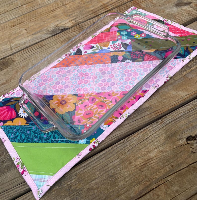 Op de afbeelding: Een heldere glazen ovenschaal ligt op een patchwork gewatteerde onderzetter. De onderzetter heeft bloemen- en geometrische patronen in roze, groen, oranje en blauw. De randen zijn afgewerkt met een roze bies. Afmetingen ca. 25 cm x 35 cm.