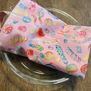 Könnte beinhalten: Ein rosa Stoffserviette mit einem bunten Süßigkeitenmuster, darunter Lutscher, Geleebohnen und Bonbons. Die Serviette ist gefaltet und liegt auf einem transparenten Glas, was einen verspielten Look erzeugt.
