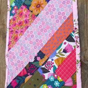 Op de afbeelding: Een kleurrijke, handgemaakte quilt met een bloemen thema. De quilt heeft diagonale strepen van verschillende patronen, waaronder bloemen, stippen en geometrische ontwerpen. De randen zijn afgewerkt met een roze bloemenrand, ongeveer 30 cm x 46 cm.