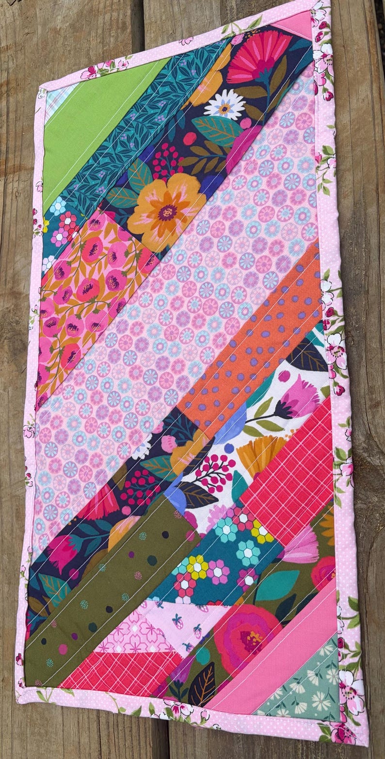 Op de afbeelding: Een kleurrijke, handgemaakte quilt met een bloemen thema. De quilt heeft diagonale strepen van verschillende patronen, waaronder bloemen, stippen en geometrische ontwerpen. De randen zijn afgewerkt met een roze bloemenrand, ongeveer 30 cm x 46 cm.