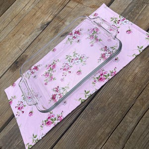 Op de afbeelding: Een heldere rechthoekige glazen ovenschaal met handvatten ligt op een roze keukendoek met bloemenpatroon. De handdoek heeft een herhalend patroon van roze en witte bloemen met groene bladeren. De achtergrond is een houten oppervlak.