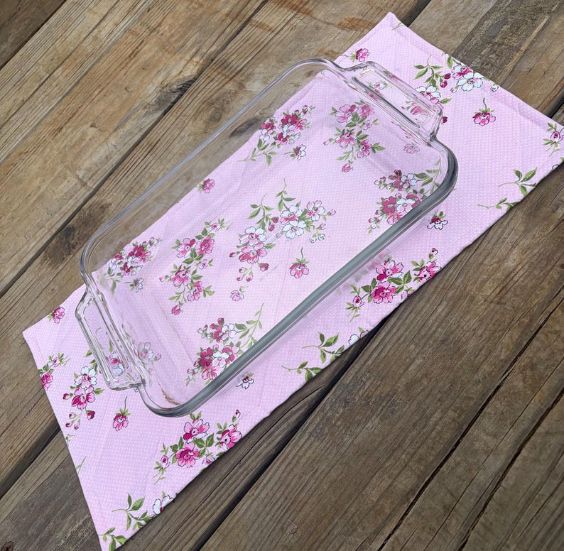 Op de afbeelding: Een heldere rechthoekige glazen ovenschaal met handvatten ligt op een roze keukendoek met bloemenpatroon. De handdoek heeft een herhalend patroon van roze en witte bloemen met groene bladeren. De achtergrond is een houten oppervlak.