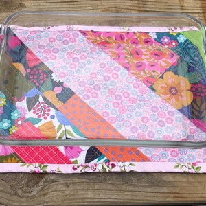 Op de afbeelding: Een heldere glazen ovenschaal rust op een kleurrijke gewatteerde mat. De mat heeft een patchwork-ontwerp met bloemen- en geometrische patronen in roze, oranje, groen en blauw. De randen van de mat zijn afgewerkt met een bloemenrand.