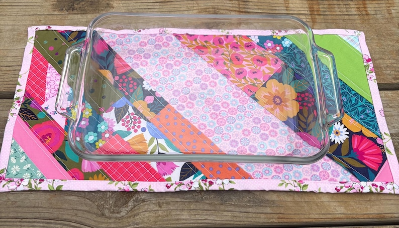 Op de afbeelding: Een heldere glazen ovenschaal rust op een kleurrijke gewatteerde mat. De mat heeft een patchwork-ontwerp met bloemen- en geometrische patronen in roze, oranje, groen en blauw. De randen van de mat zijn afgewerkt met een bloemenrand.