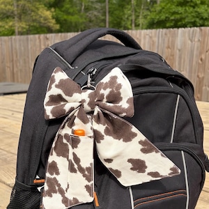 May include: A black backpack with a cow print bow-shaped keychain attached. The bow is white with brown spots. The backpack has orange accents and a small pocket. The background is a wooden deck and trees.