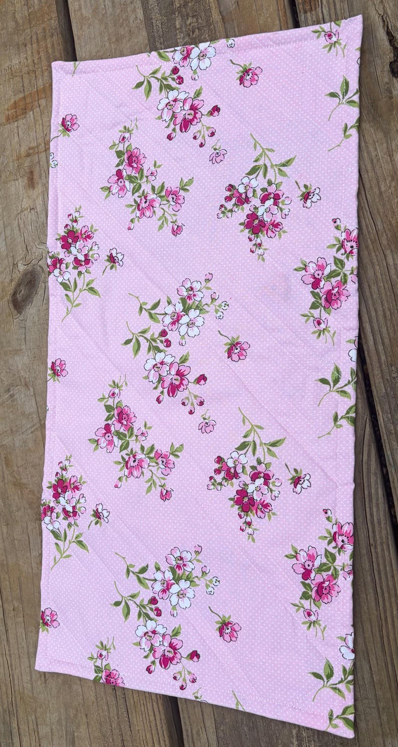 Op de afbeelding: Roze bloemenstof met witte stippen. De stof heeft een herhalend patroon van roze en witte bloemen met groene bladeren. De stof is gewatteerd met diagonale stiksels.