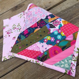 Op de afbeelding: Een handgemaakte quilt met een patchwork-ontwerp. De quilt toont een verscheidenheid aan bloemen- en geometrische patronen in roze, groene, blauwe en rode tinten. De randen zijn afgewerkt met een bloemenprint, waardoor een decoratief item ontstaat.