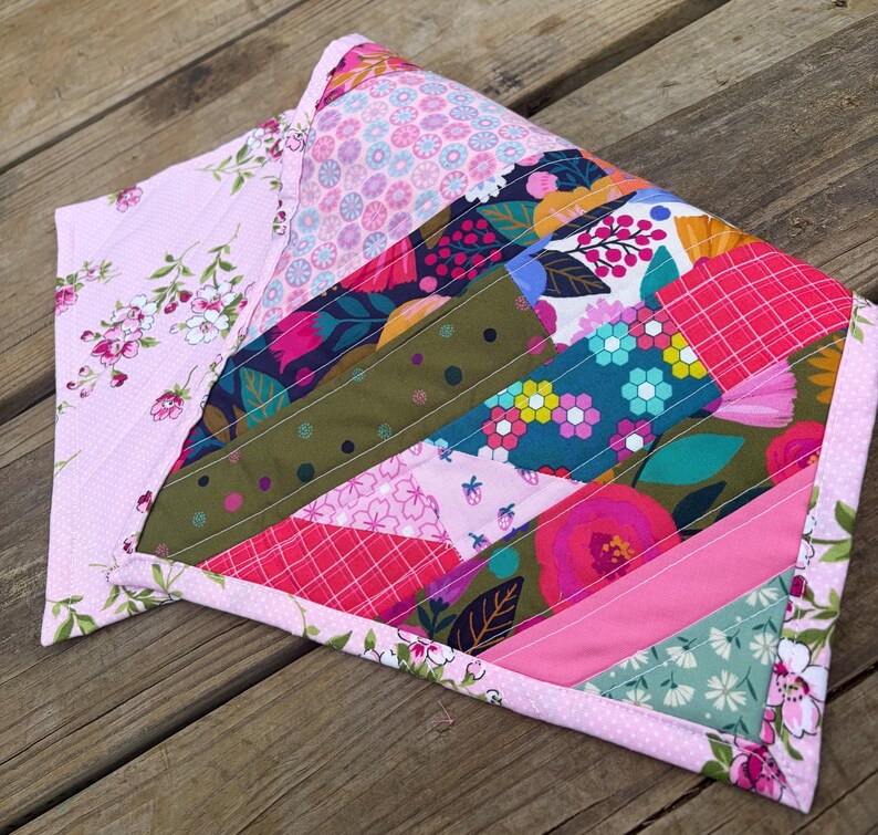 Op de afbeelding: Een handgemaakte quilt met een patchwork-ontwerp. De quilt toont een verscheidenheid aan bloemen- en geometrische patronen in roze, groene, blauwe en rode tinten. De randen zijn afgewerkt met een bloemenprint, waardoor een decoratief item ontstaat.