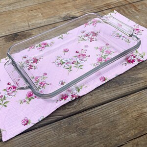 Op de afbeelding: Een rechthoekige glazen ovenschaal met handvatten ligt op een roze keukendoek met bloemenpatroon. De handdoek heeft een herhalend patroon van roze en witte bloemen met groene bladeren. De achtergrond is hout.