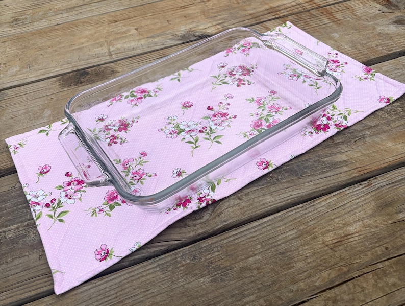 Op de afbeelding: Een rechthoekige glazen ovenschaal met handvatten ligt op een roze keukendoek met bloemenpatroon. De handdoek heeft een herhalend patroon van roze en witte bloemen met groene bladeren. De achtergrond is hout.
