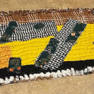 May include: A handmade, rectangular rag rug with a patchwork design. The rug features a combination of fabrics in various colors and patterns, including brown plaid, black and white gingham, yellow, and orange. The edges are fringed.