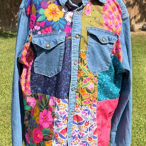 Könnte beinhalten: Jeansjacke mit farbenfrohen Blumen-Patchwork-Einsätzen. Die Jacke hat zwei Brusttaschen und eine Knopfleiste. Der Patchwork-Stil umfasst leuchtende rosa, gelbe und blaue Blumenmuster und schafft ein einzigartiges und auffälliges Design.