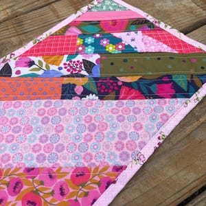 Op de afbeelding: Een kleurrijke, gewatteerde tafelloper met een patchwork-ontwerp. De loper heeft verschillende bloemen- en geometrische patronen in roze, groene en blauwe tinten. De randen zijn afgewerkt met een roze bloemenrand.