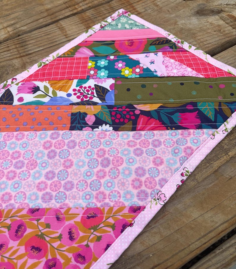 Op de afbeelding: Een kleurrijke, gewatteerde tafelloper met een patchwork-ontwerp. De loper heeft verschillende bloemen- en geometrische patronen in roze, groene en blauwe tinten. De randen zijn afgewerkt met een roze bloemenrand.