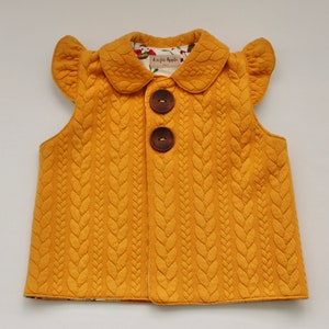 May include: A mustard yellow sleeveless vest with a cable knit pattern and a white Peter Pan collar. The vest has two brown buttons and ruffled sleeves.