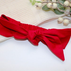 Könnte beinhalten: Ein rotes Stoffhaarband, das zu einer Schleife gebunden ist. Das Haarband liegt auf einer weißen gestrickten Oberfläche.