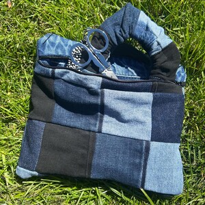 May include: A patchwork denim handbag with a zip closure and a rounded handle. The bag is made from a mix of blue and black denim squares. A pair of silver scissors rests on top of the bag, set against green grass.