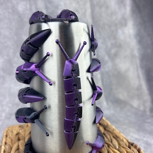 Peut inclure: Un gobelet en acier inoxydable avec un motif de escargots violet et noir. Les escargots sont disposés autour du gobelet, avec un gros escargot au centre.