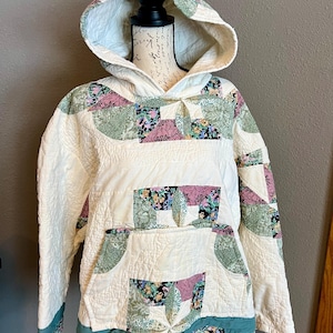 May include: Cream-colored quilted hoodie with a patchwork design. The hoodie features a front pocket and a hood with floral and geometric patterns in shades of pink, green, and blue. The bottom hem and sleeve cuffs have a teal floral trim.
