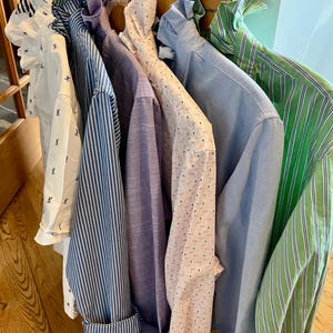 May include: A collection of button-down shirts in various colors and patterns hanging on wooden hangers. The shirts include white with black accents, blue stripes, solid lavender, pink with small dots, light blue, and green stripes.