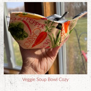 May include: A beige fabric bowl cozy with a black and white checkered trim. The cozy features a colorful print of carrots, peppers, and other vegetables with the words "Chili Pepper" and "Carrot" printed on the fabric. The cozy is being held by a hand in front of a window.