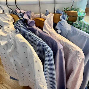 May include: A row of long-sleeved button-down shirts hanging on wooden hangers. The shirts feature various patterns including small black skull motifs on white, blue and white vertical stripes, solid light purple, pink with dots, and green stripes.