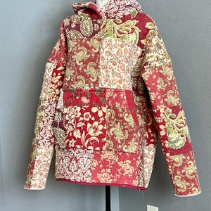 May include: A quilted hoodie in a patchwork design, featuring a mix of floral and paisley patterns in shades of red, cream, and green. The hoodie has a front pocket and a hood, displayed on a mannequin.