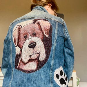 May include: A blue denim jacket featuring a large, detailed embroidered patch of a brown and white dog on the back. Below the dog patch, there is a smaller, circular embroidered patch depicting a black paw print on a white background.