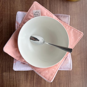 May include: A white ceramic bowl and a silver spoon rest on two pink and white fabric pot holders. The top pot holder has a pink background with white polka dots. The bottom pot holder has a pink and white striped pattern. The top pot holder has a tag that says "jolly design co handmade".