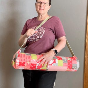 May include: A colorful yoga mat bag with a patchwork design in shades of red, pink, green, and gray. The bag has a long, light green strap for carrying. The bag is cylindrical and appears to be made of fabric.