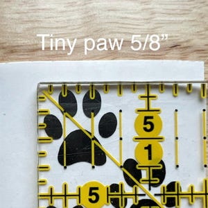 Puede incluir: Un gr&aacute;fico de huella de pata negra sobre una superficie blanca, parcialmente cubierto por una regla de acr&iacute;lico transparente con marcas amarillas. El texto "Tiny paw 5/8"" indica que la huella mide 1,59 cm de tama&ntilde;o.