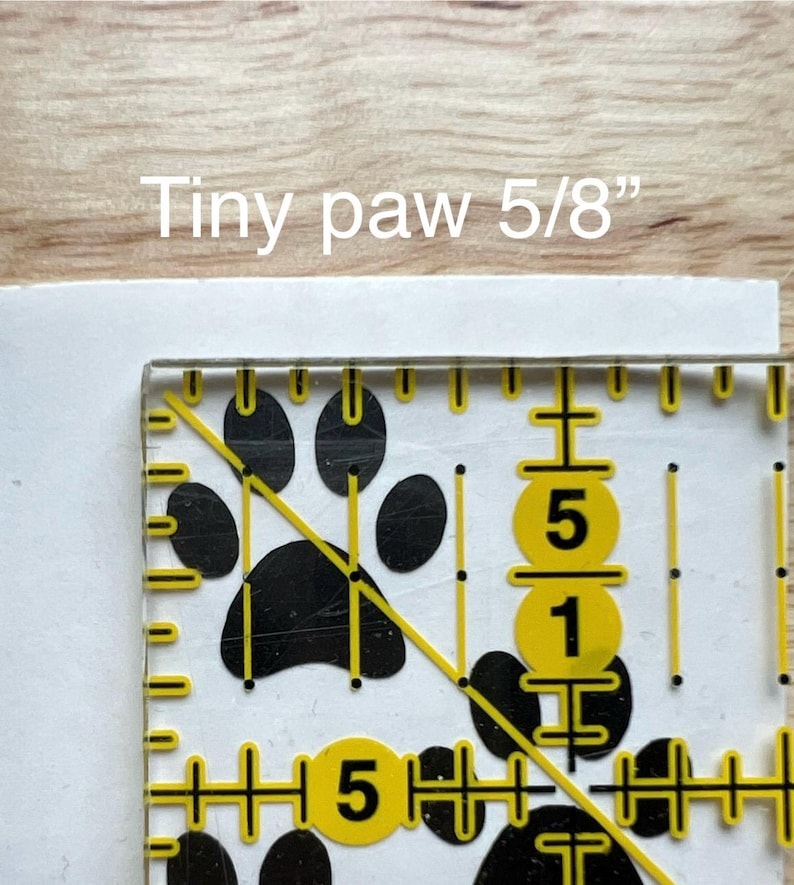 Puede incluir: Un gr&aacute;fico de huella de pata negra sobre una superficie blanca, parcialmente cubierto por una regla de acr&iacute;lico transparente con marcas amarillas. El texto "Tiny paw 5/8"" indica que la huella mide 1,59 cm de tama&ntilde;o.