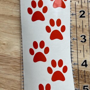 Puede incluir: Una hoja de pegatinas blanca vertical con siete huellas de pata rojas dispuestas en columna. El texto "Small" est&aacute; impreso en la parte superior. Una regla transparente a la derecha muestra que la hoja mide unos 15 cm de largo.