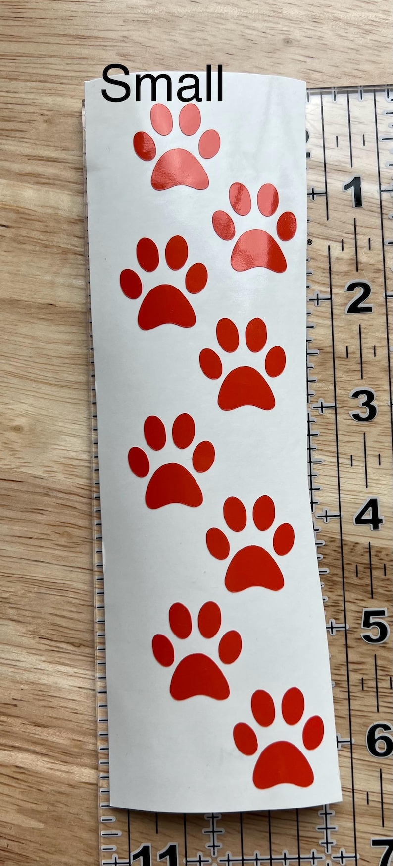 Puede incluir: Una hoja de pegatinas blanca vertical con siete huellas de pata rojas dispuestas en columna. El texto "Small" est&aacute; impreso en la parte superior. Una regla transparente a la derecha muestra que la hoja mide unos 15 cm de largo.