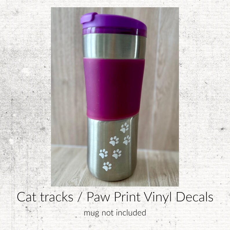 Puede incluir: Una taza de viaje de acero inoxidable con tapa morada y una banda de agarre de silicona morada ancha alrededor del centro. La parte inferior de la taza est&aacute; decorada con varias pegatinas blancas peque&ntilde;as de huellas de patas.