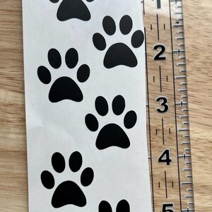 Puede incluir: Una hoja de pegatinas blanca con cinco huellas de patas negras dispuestas verticalmente. La palabra Large est&aacute; impresa en la parte superior. Una regla de pl&aacute;stico transparente a la derecha muestra que la hoja mide unos 15 cm.