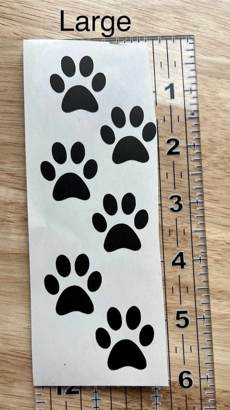 Puede incluir: Una hoja de pegatinas blanca con cinco huellas de patas negras dispuestas verticalmente. La palabra Large est&aacute; impresa en la parte superior. Una regla de pl&aacute;stico transparente a la derecha muestra que la hoja mide unos 15 cm.