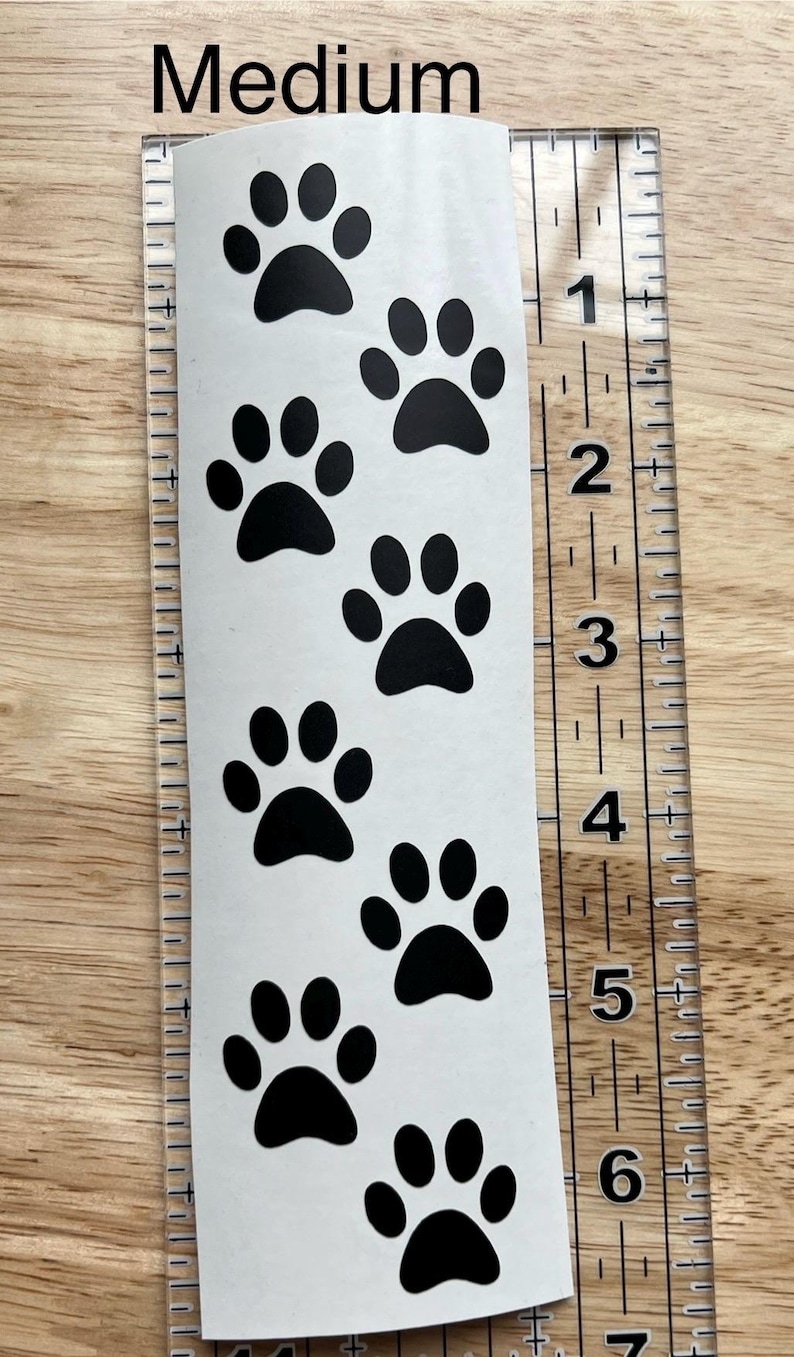 Puede incluir: Una calcoman&iacute;a blanca de tama&ntilde;o mediano con un patr&oacute;n vertical de huellas de patas negras, colocada junto a una regla de pl&aacute;stico transparente que muestra que la calcoman&iacute;a mide aproximadamente 16,5 cm de largo. La palabra Medium est&aacute; impresa arriba.