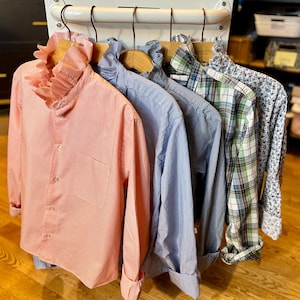 May include: A row of five long-sleeved button-down shirts hanging on wooden hangers. The shirts feature various patterns including pink pinstripes with a ruffled collar, blue pinstripes, blue gingham, green and blue plaid, and a blue floral print.