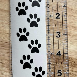 Puede incluir: Una tira adhesiva blanca con una serie de huellas de patas negras dispuestas en un sendero vertical. La palabra Tiny est&aacute; impresa en la parte superior. Una regla de pl&aacute;stico transparente est&aacute; junto al adhesivo, mostrando 16,5 cent&iacute;metros.