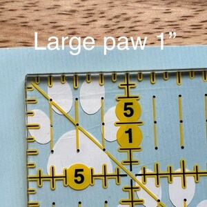 Puede incluir: Una superficie azul claro con un patr&oacute;n de huellas blancas, superpuesta con una regla de acolchado transparente marcada con l&iacute;neas y n&uacute;meros amarillos. El texto "Large paw 1"" indica el tama&ntilde;o de la huella.
