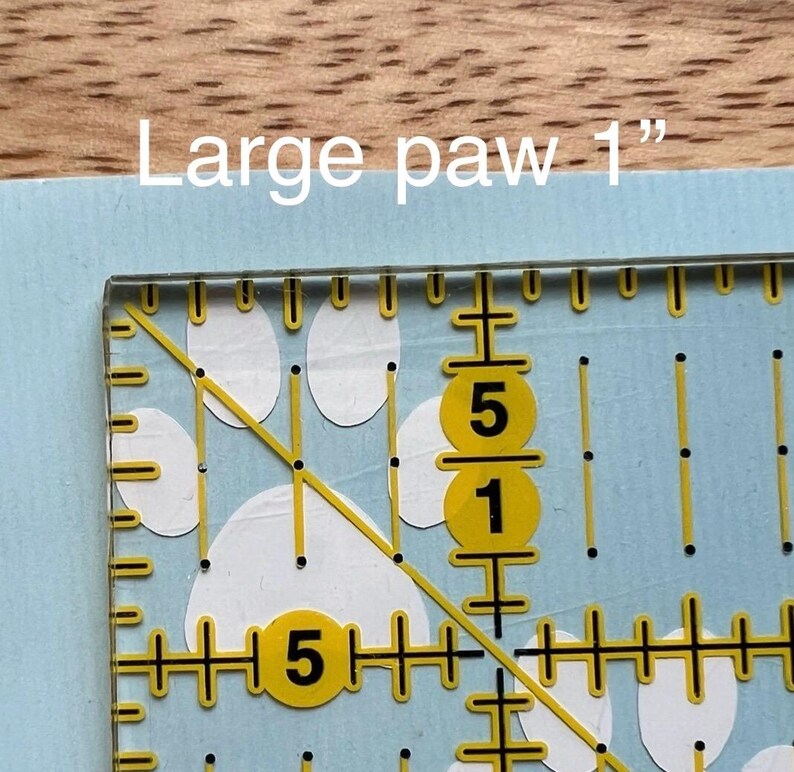 Puede incluir: Una superficie azul claro con un patr&oacute;n de huellas blancas, superpuesta con una regla de acolchado transparente marcada con l&iacute;neas y n&uacute;meros amarillos. El texto "Large paw 1"" indica el tama&ntilde;o de la huella.