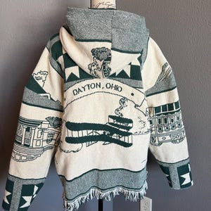 May include: Cream-colored hooded jacket with green accents and fringe. The jacket features a design with the text "DAYTON, OHIO" and illustrations of buildings and airplanes. The hood and trim are green, and the jacket is displayed on a mannequin.