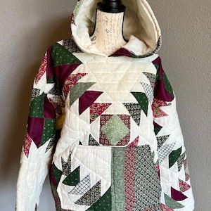 May include: A quilted hoodie in a patchwork design. The hoodie features a cream-colored base with geometric patterns in green, burgundy, and patterned fabrics. It has a hood, a front pocket, and long sleeves. The garment is displayed on a mannequin.