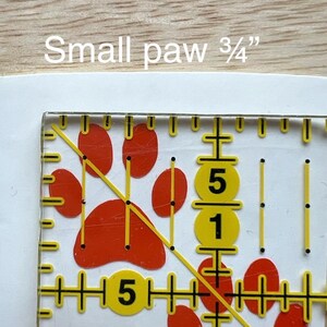 Puede incluir: Una regla de acolchado de acr&iacute;lico transparente con un dise&ntilde;o de huella de pata roja y marcas de medici&oacute;n amarillas. El texto "Small paw 3/4"" es visible en la parte superior. La regla incluye una l&iacute;nea diagonal e indicadores num&eacute;ricos.