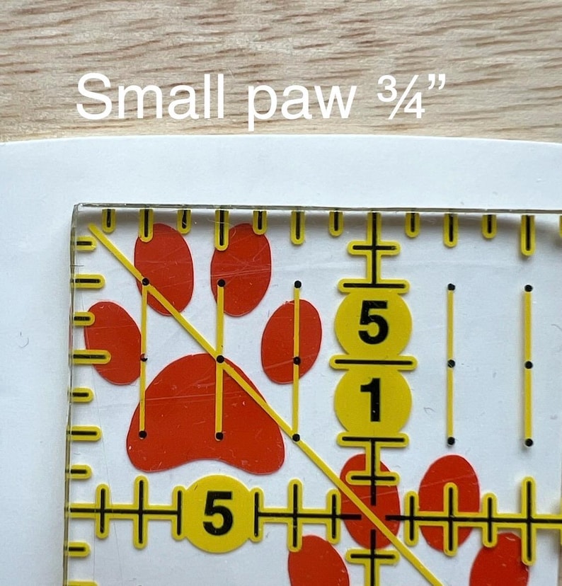 Puede incluir: Una regla de acolchado de acr&iacute;lico transparente con un dise&ntilde;o de huella de pata roja y marcas de medici&oacute;n amarillas. El texto "Small paw 3/4"" es visible en la parte superior. La regla incluye una l&iacute;nea diagonal e indicadores num&eacute;ricos.