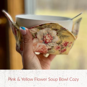 May include: A white ceramic soup bowl with a spoon inside, held by a pink and yellow floral soup bowl cozy. The cozy features a vintage floral pattern with pink and red flowers on a yellow background. The text at the bottom reads "Pink & Yellow Flower Soup Bowl Cozy."