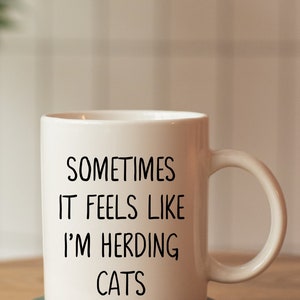 Może przedstawiać: Biały ceramiczny kubek z czarnym napisem "Sometimes it feels like I'm herding cats". Kubek stoi na zielonym marmurowym podkładce.