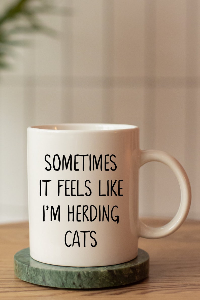 Może przedstawiać: Biały ceramiczny kubek z czarnym napisem "Sometimes it feels like I'm herding cats". Kubek stoi na zielonym marmurowym podkładce.