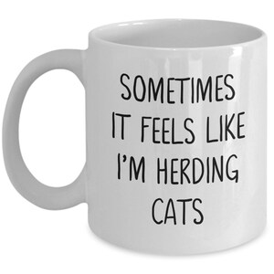 Może przedstawiać: Biały ceramiczny kubek do kawy z czarnym tekstem, kt&oacute;ry brzmi "Sometimes it feels like I'm herding cats".